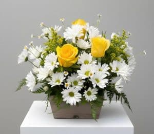 Bouquet of yellow roses and white daisies in a rectangular wooden planter on a white pedestal.