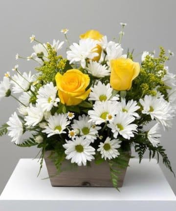 Bouquet of yellow roses and white daisies in a rectangular wooden planter on a white pedestal.
