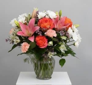 A glass vase on a white table holds a bright bouquet of pink lilies, orange roses, peach carnations, and white flowers with greenery.