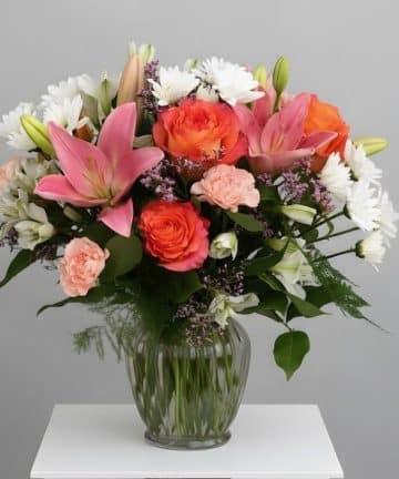 A glass vase on a white table holds a bright bouquet of pink lilies, orange roses, peach carnations, and white flowers with greenery.