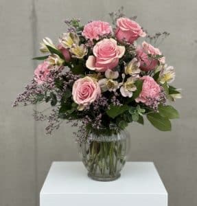 Pink bouquet with roses and alstroemeria in a glass vase on a white pedestal, with greenery and baby's breath accents.