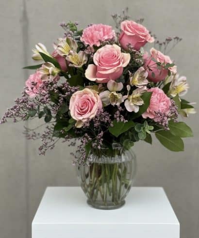 Pink bouquet with roses and alstroemeria in a glass vase on a white pedestal, with greenery and baby's breath accents.